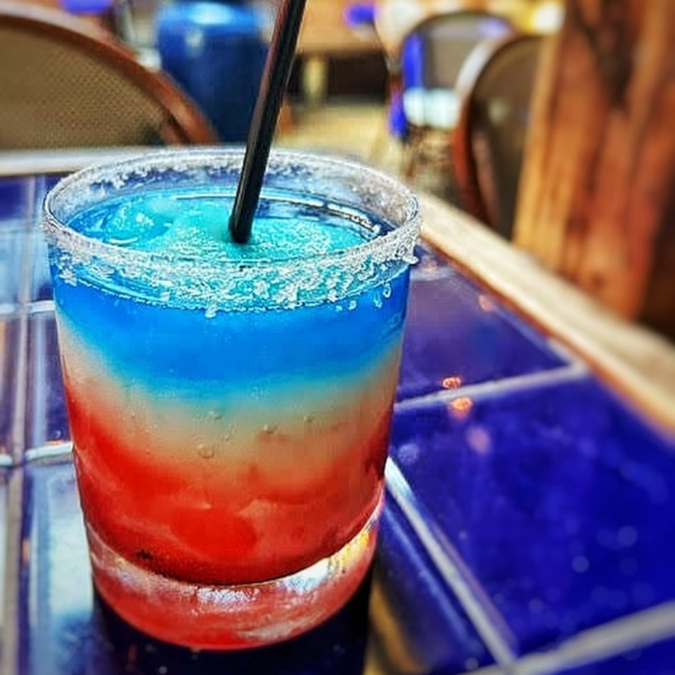 Red, white, and blue layered frozen drink with a straw and salt rim on a blue tiled table.